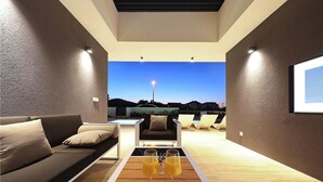 Un salon moderne comprenant un canapé, deux fauteuils, une table basse avec des boissons, et une grande fenêtre offrant une vue sur le ciel et les bâtiments éloignés.