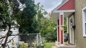 Un jardin arrière d’une maison, avec un chemin en béton menant à un porche, le tout entouré de verdure et d’une clôture.