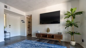 Una sala moderna con un televisor de pantalla plana grande, un mueble de entretenimiento de madera, una planta grande en maceta y una alfombra azul estampada.