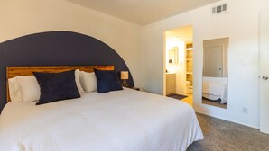 Una cama bien tendida con cabecera de madera, un espejo grande en la pared y un baño visible a través de una puerta abierta.
