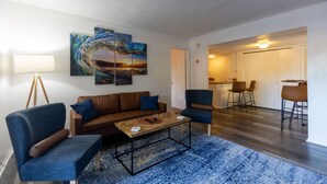 Una sala de estar con un sofá marrón, sillones azules, una mesa de centro y un tapete azul. Hay tres cuadros en la pared, una lámpara y una zona de comedor al fondo.