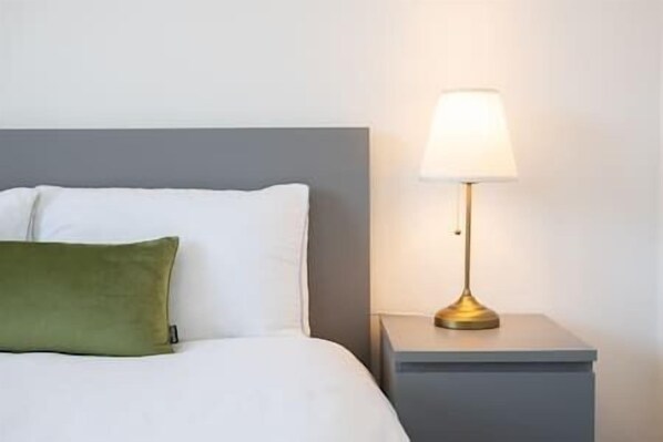A bed with white bedding and a green pillow, a bedside table with a lamp, and a grey nightstand.