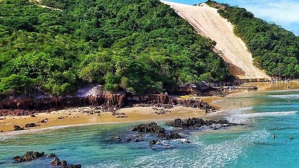 A beach with clear turquoise water, rocky shore, and a forested hill. - Encantadora casa em Ponta Negra (Natal)
