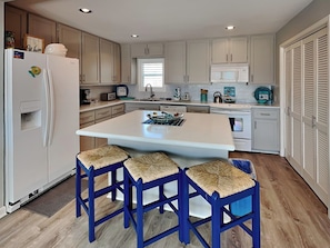 A modern kitchen with white cabinets, a central island with stools, and a refrigerator with magnets. - Pirates' View of the Sea by Ryson Vacation Rentals (Galveston)