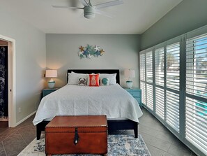 Un dormitorio con cama, mesitas de noche, cómoda, ventilador de techo y una ventana grande con persianas.