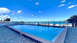A rectangular swimming pool with a tiled deck, a white fence, and lounge chairs along the poolside. - Oceanside Retreat 804 by Distinctive Beach Rentals (Ormond Beach)