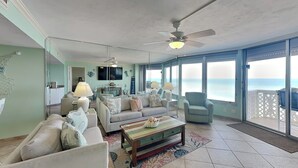 A spacious living room with a sofa set, a coffee table, and a television. There is a balcony with a view of the ocean. - Oceanside Retreat 804 by Distinctive Beach Rentals (Ormond Beach)