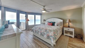 A bedroom with a bed, a nightstand, a lamp, a ceiling fan, and a view of the ocean. - Oceanside Retreat 804 by Distinctive Beach Rentals (Ormond Beach)