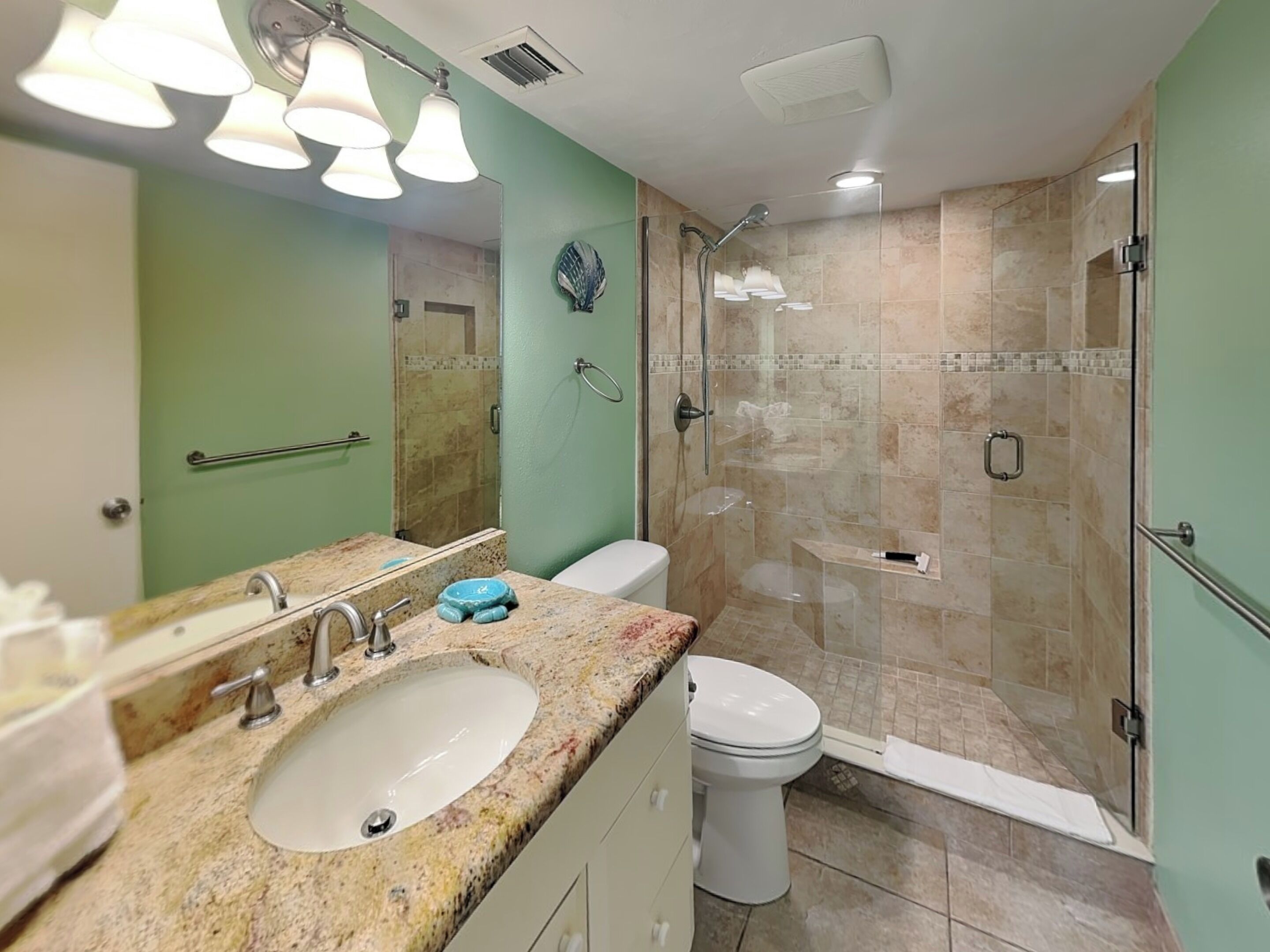 A bathroom with a granite countertop sink, a white toilet, a glass-enclosed shower, and a mirror with three lights.