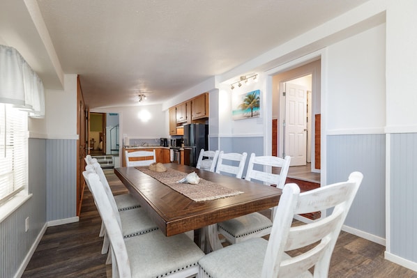 A dining area with a wooden table and white chairs, a kitchen in the background, and a door leading to another room.