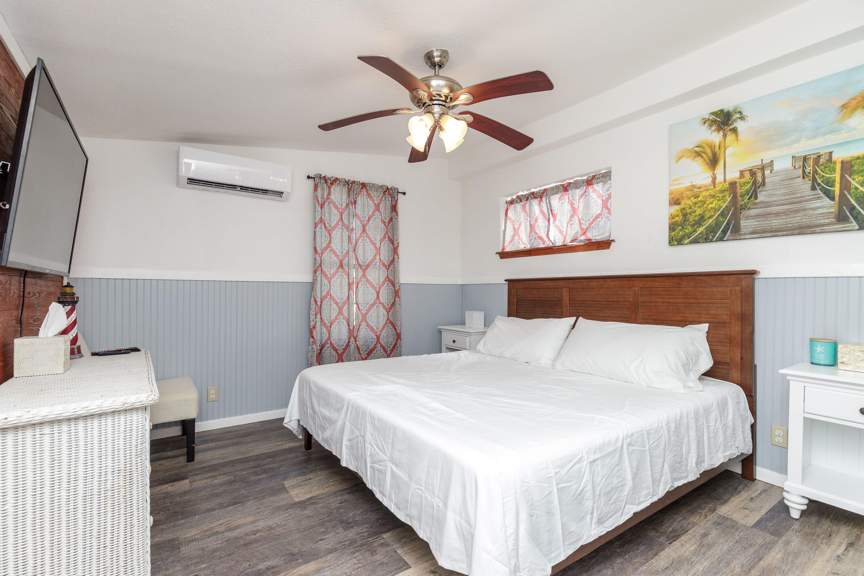 Une chambre à coucher comprenant un lit, un ventilateur de plafond, un climatiseur, une table de chevet, une lampe, un tableau et une fenêtre avec des rideaux.