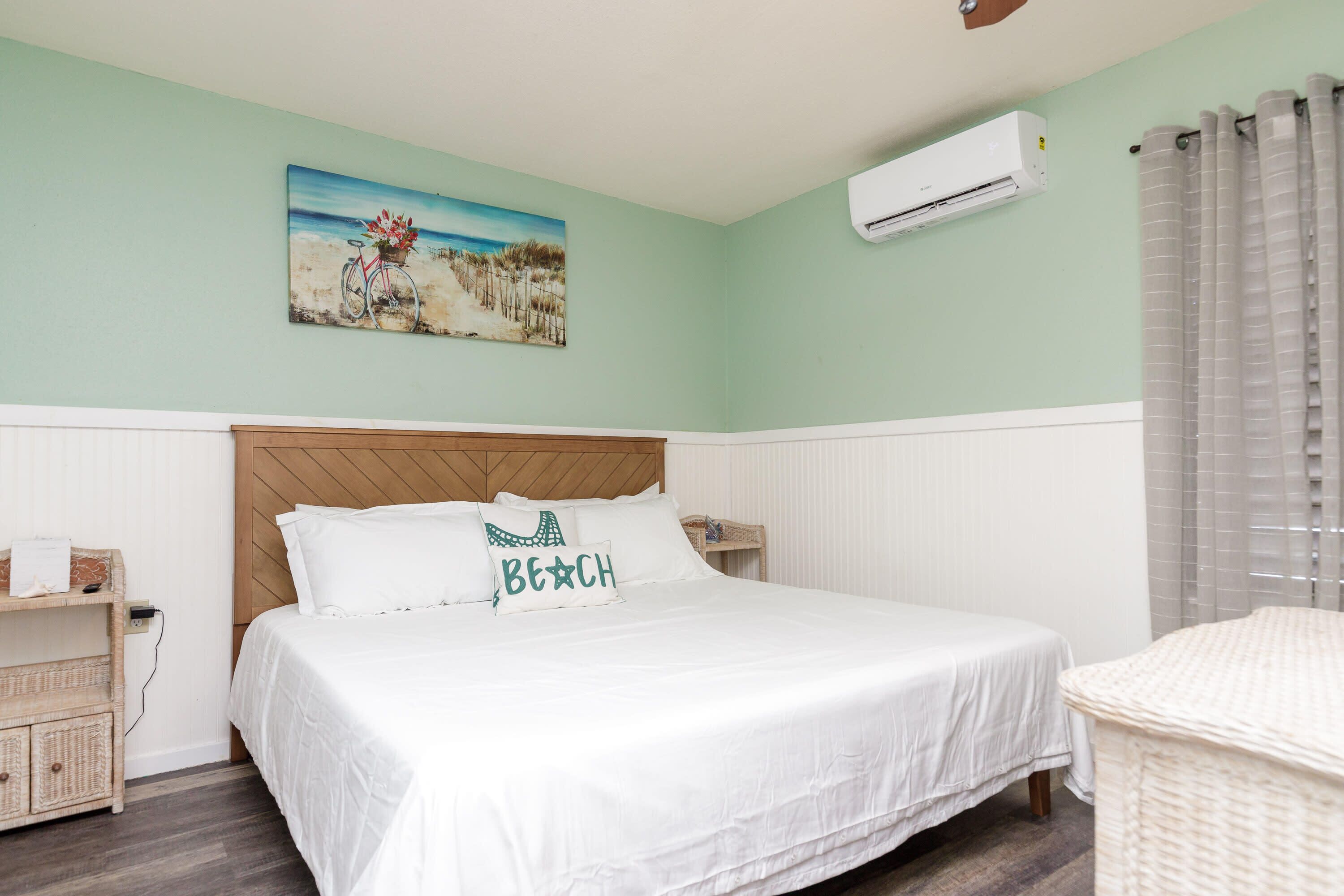 Ein Schlafzimmer mit einem Bett, einem Kopfteil, einer Wand-Klimaanlage, einem Bild mit Strandmotiv und einem Weiden-Nachttisch.