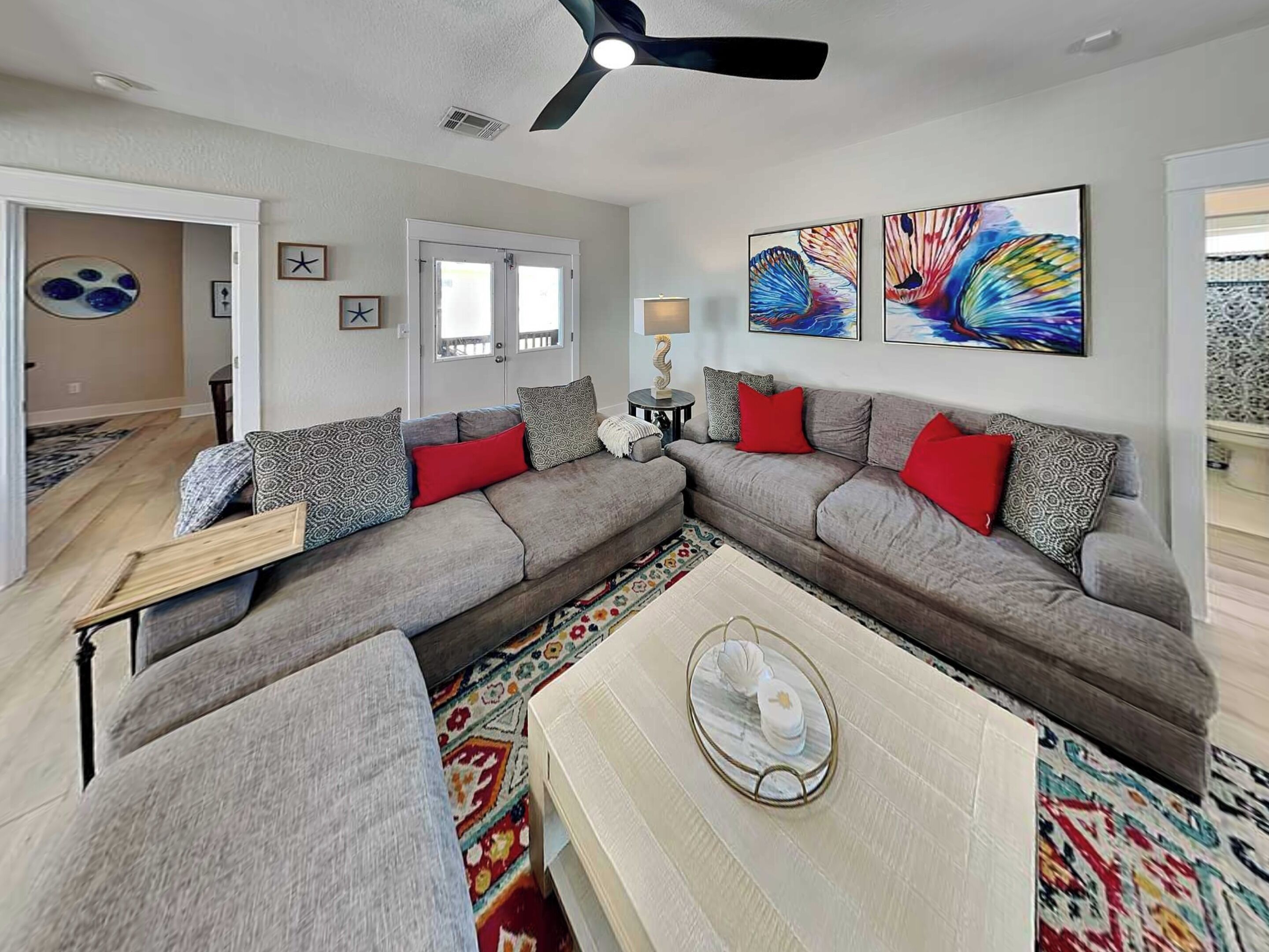 Un salon moderne avec un canapé d’angle gris, une table basse et un tapis coloré. Deux tableaux ornent le mur, ainsi qu’un ventilateur de plafond et une porte qui donne sur une autre pièce.