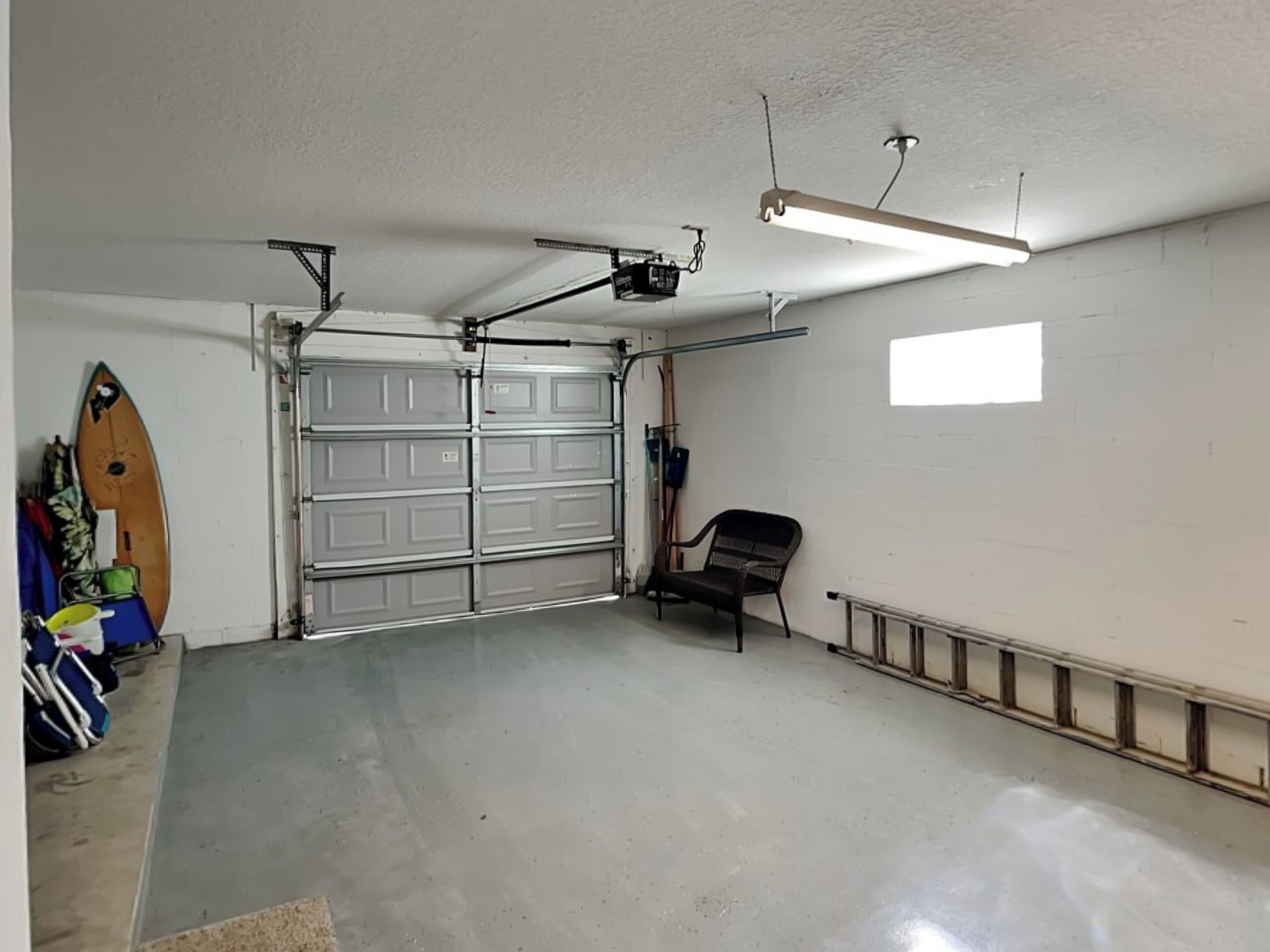 Eine Garage mit Betonboden, zwei Garagentoren, einem an der Decke befestigten Projektor und einem Surfbrett, das an der Wand lehnt.