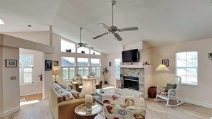 A spacious living room with a fireplace, ceiling fans, and a television. - Seascape Beachhouse by Distinctive Beach Rentals (St. Augustine)