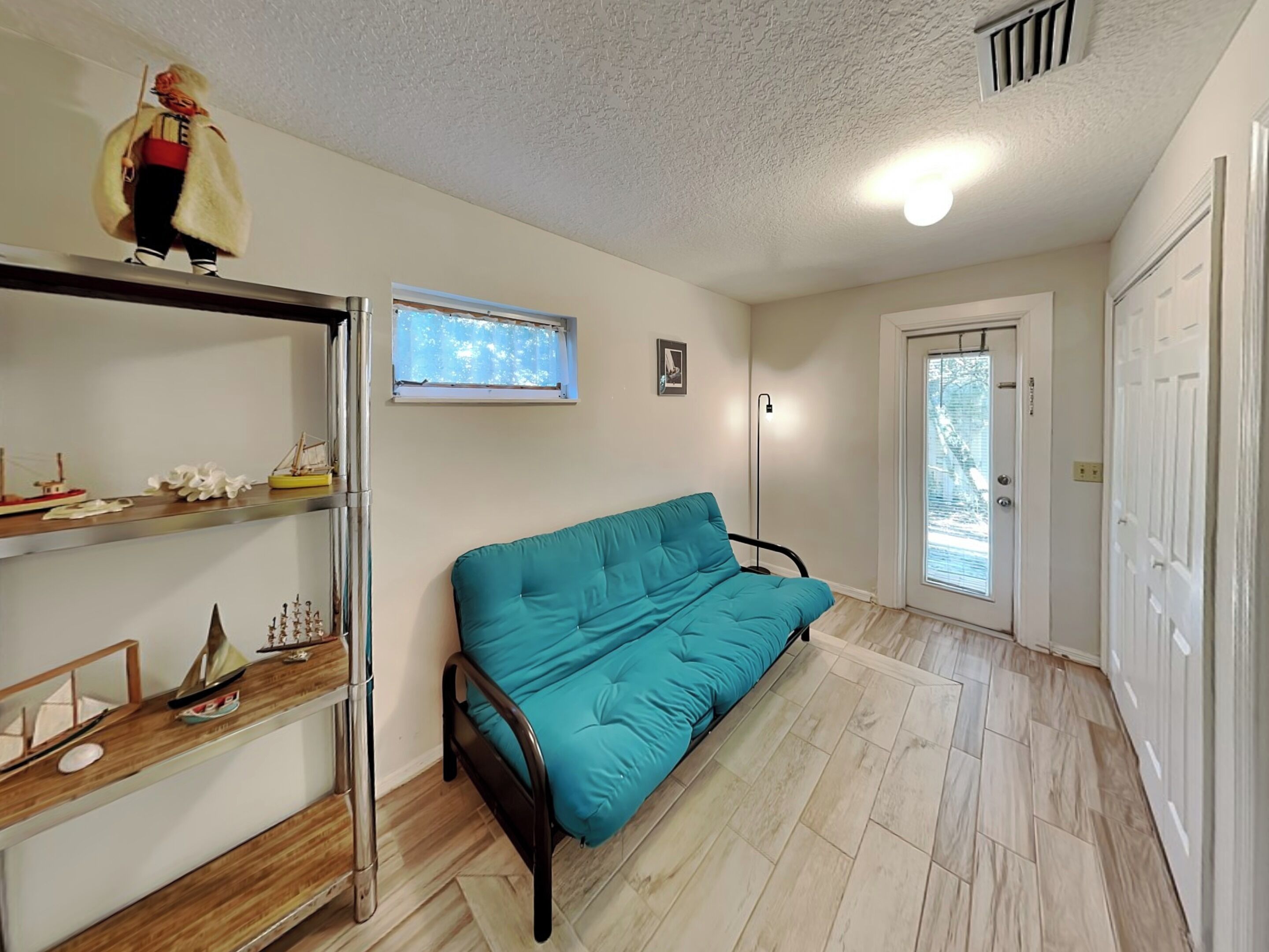 A room with a teal sofa, a wooden shelf with model ships, a window with blinds, a door, and a light fixture.