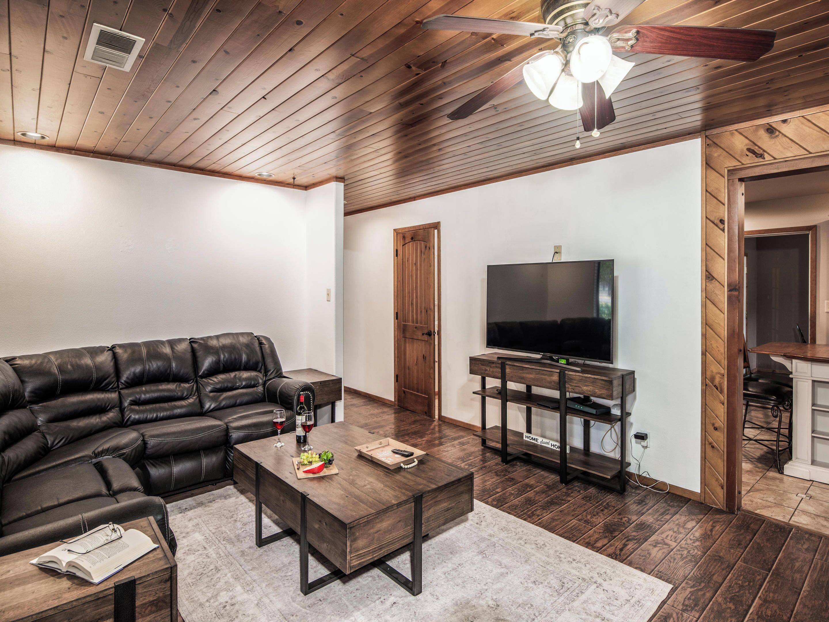 A living room with a leather sofa, a wooden coffee table, a flat-screen TV on a wooden stand, and a ceiling fan.