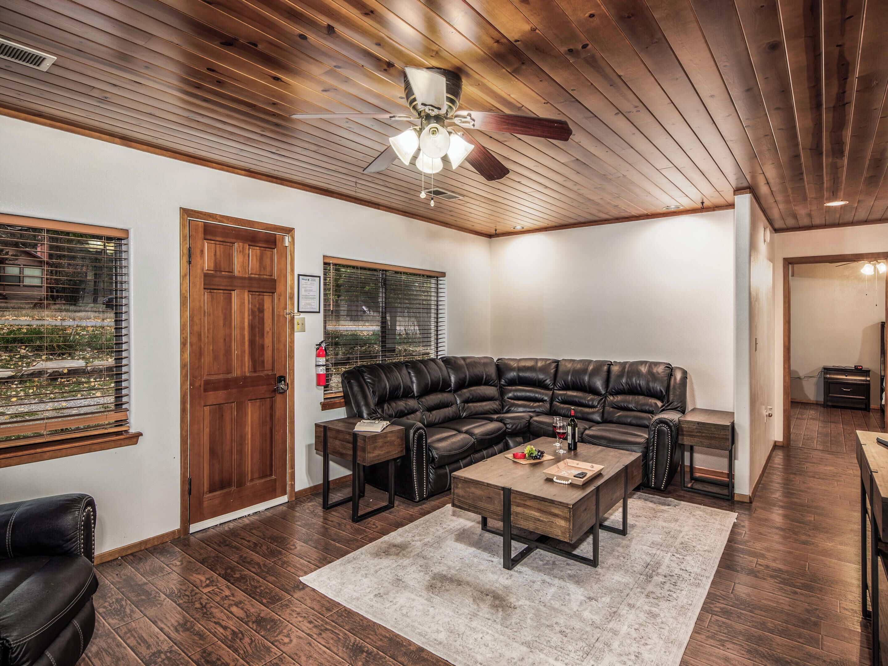 A living room with a sectional sofa, a coffee table, wooden flooring, a ceiling fan, and a window with blinds.