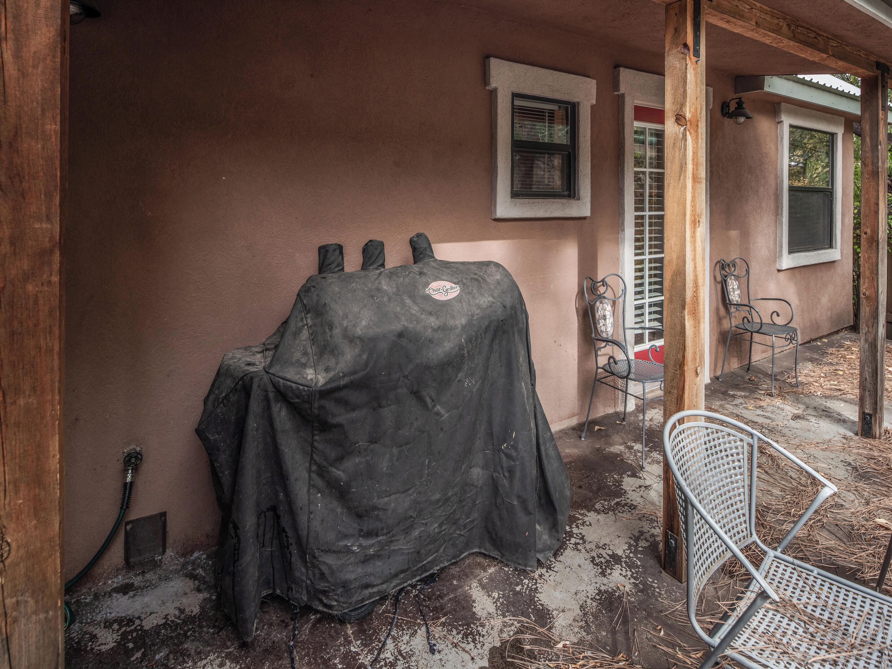 A covered grill area with a hose, a metal chair, and a bench under a porch.