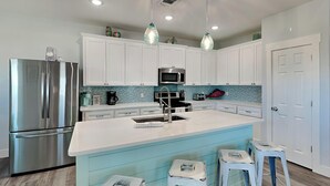 A modern kitchen with white cabinets, a stainless steel refrigerator, a microwave, and a large island with stools. - Mambo's Grill by Miss Kitty’s (Rockport)