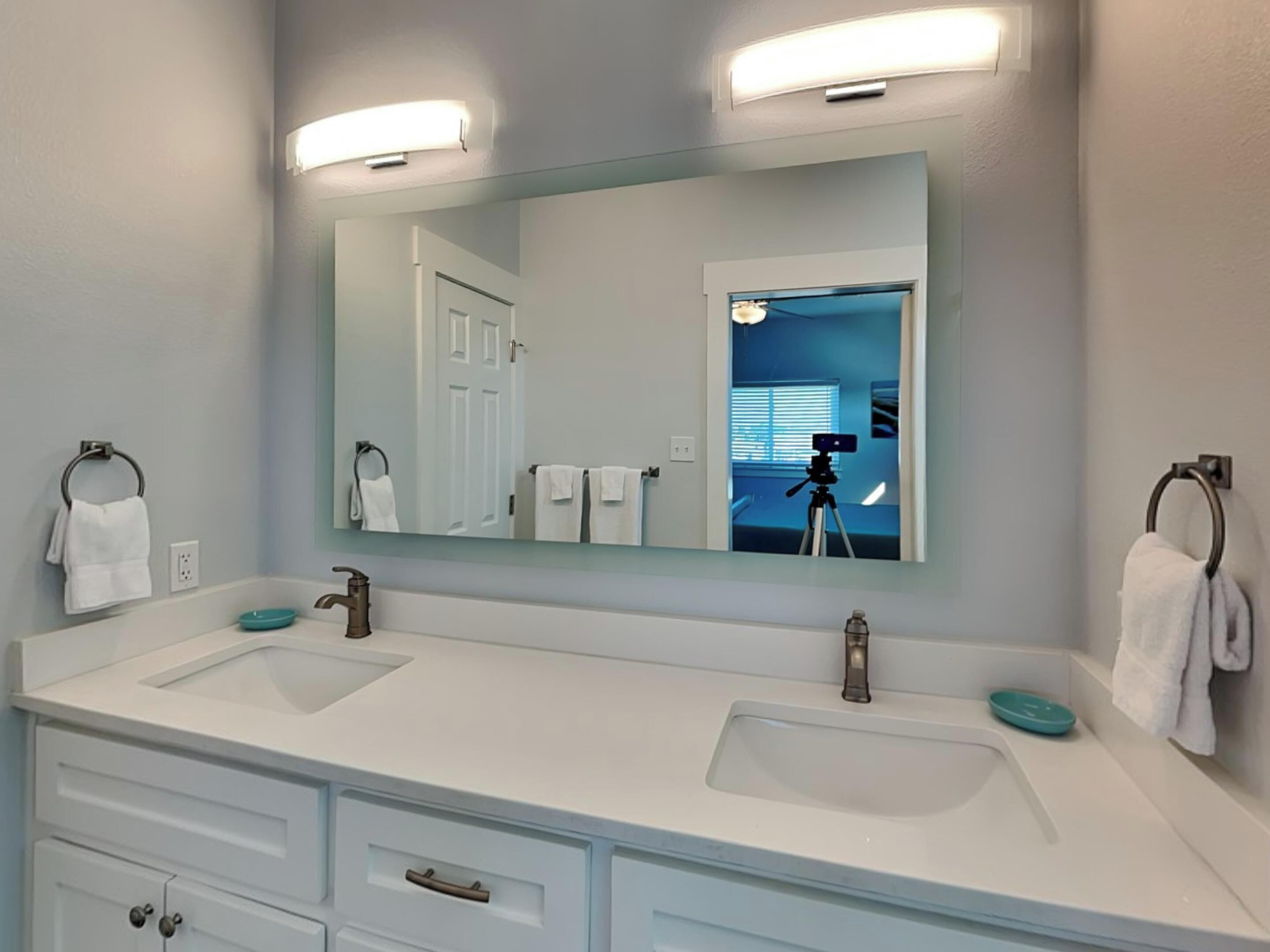 A bathroom with two sinks, a large mirror, and a camera on a tripod in the reflection.