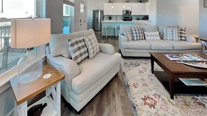 Una sala de estar moderna con un sofá, una mesa de centro y una lámpara. Se observa una cocina al fondo con un refrigerador y un microondas.