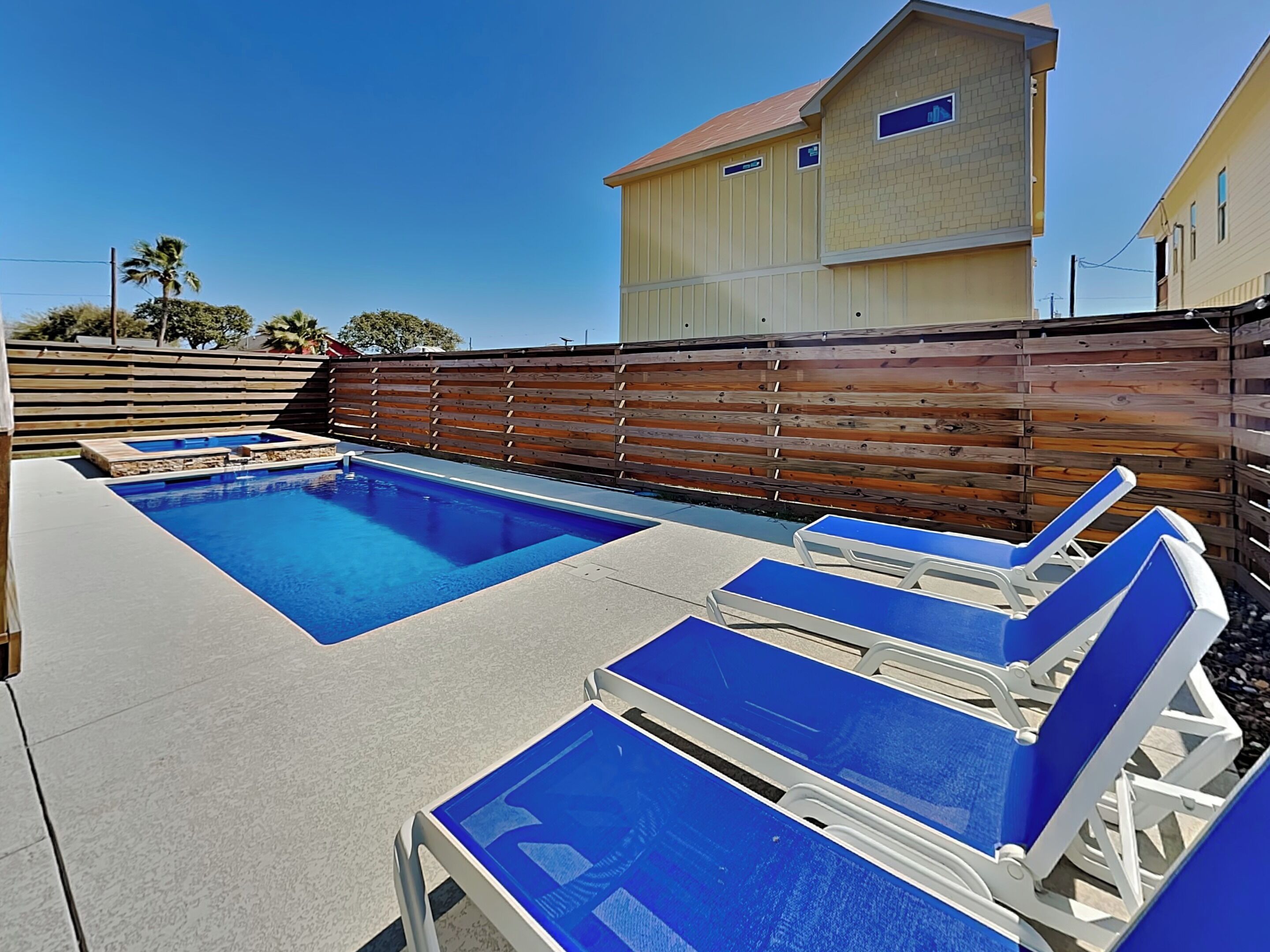 A swimming pool with lounge chairs, a wooden fence, and a two-story building in the background.