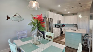 A modern kitchen with white cabinets, a central island, and a dining area with a wooden table and teal chairs. - Mambo's Grill by Miss Kitty’s (Rockport)