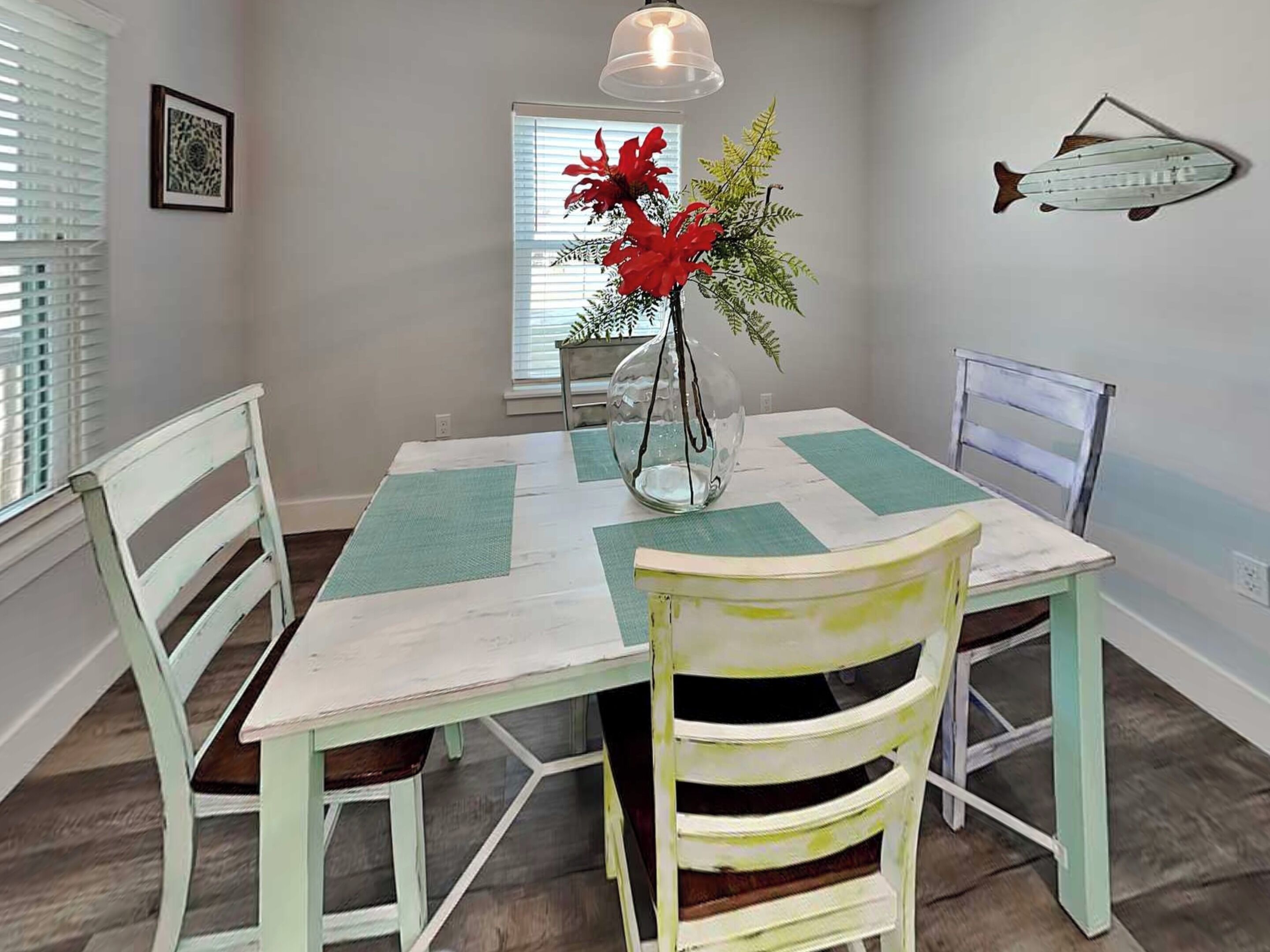 Uma área de refeições com mesa, cadeiras, um vaso com flores, um adorno de peixe fixado na parede e uma janela com persianas.