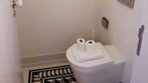 Kylpyhuoneessa on wc, kaksi rullaa wc-paperia, pyyhe, mustavalkoinen kuvioitu matto ja kolme kehystettyä kuvaa seinällä.