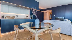 Eine moderne Küche mit integrierter Kochinsel, einem Essbereich mit Glastisch und Holzstühlen sowie einem Wohnbereich mit Sofa und Fernseher.