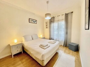 Ein Schlafzimmer mit einem Bett, Nachttischen, einem kleinen grauen Mülleimer und einem Fenster mit Vorhängen.