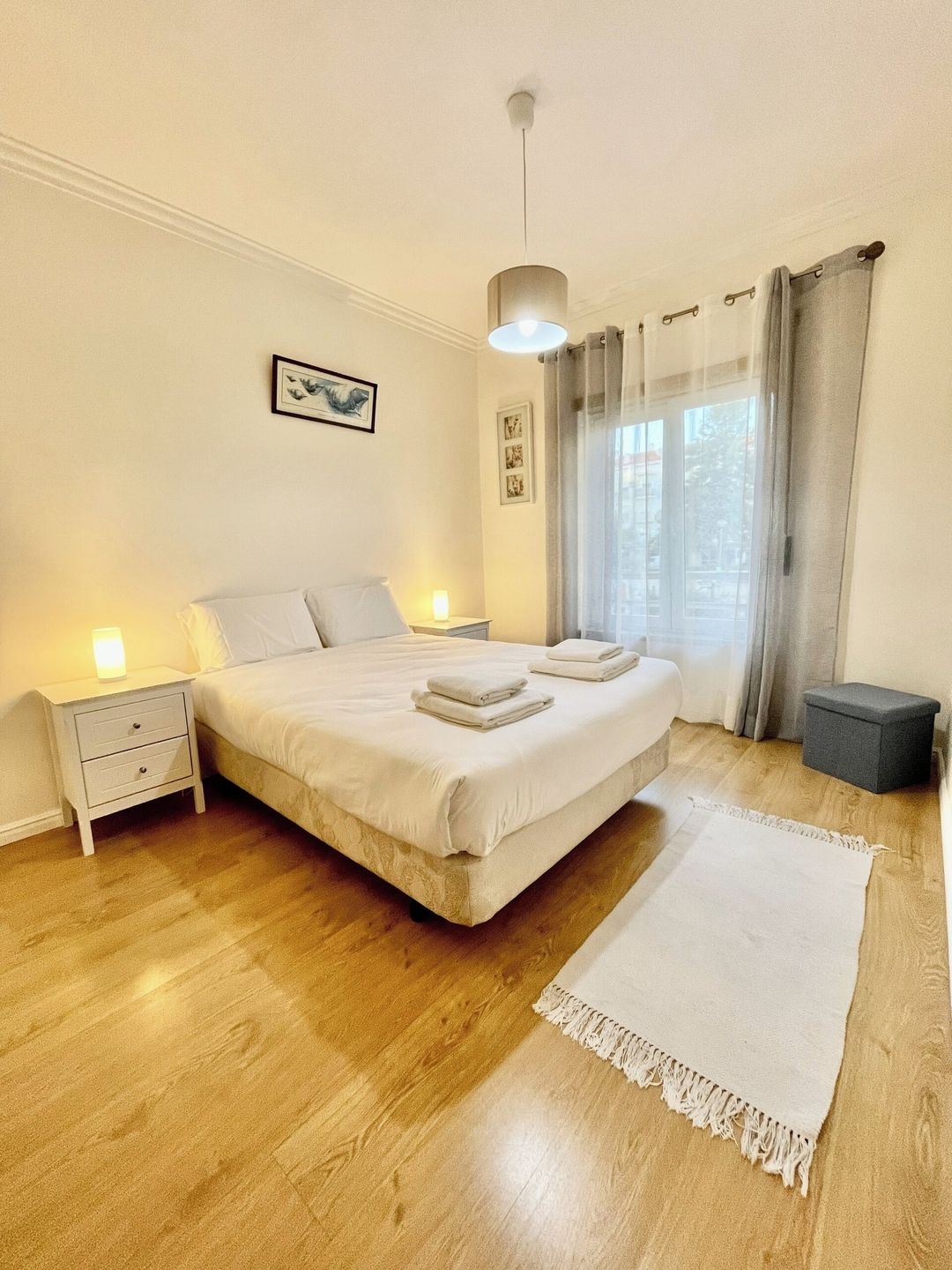 A bedroom with a large bed, bedside tables, a hanging light fixture, a window with curtains, and a small stool.