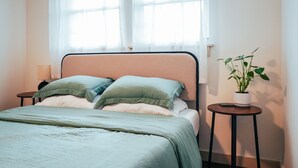 Un lit bien fait, avec une tête de lit de couleur pêche, une table de chevet sur laquelle se trouve une lampe, et une plante en pot posée sur une petite table à côté du lit.