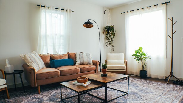 Un salon avec un canapé en cuir marron, une table basse, un lampadaire, un fauteuil et une grande fenêtre avec des rideaux.