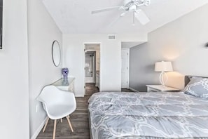 Quarto com cama, escrivaninha, cadeira, ventilador de teto e mesa de cabeceira com abajur.
