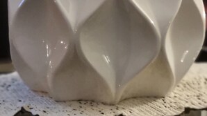 Un vase en céramique blanche à la surface texturée, orné d’un motif en forme de feuille sur le dessus, posé sur un chemin de table en dentelle.