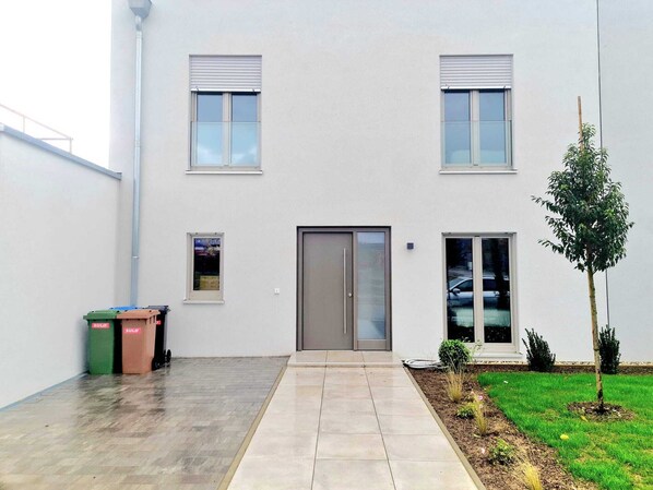 A modern building with a flat facade, a central entrance with a glass door, and a small garden area with a tree and shrubs.