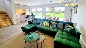 A modern living room with a green sectional sofa, a glass coffee table, and a dining area with a view of the outdoors. - Spacious & Luxurious 5 bedrooms House (Reckange-sur-Mess)