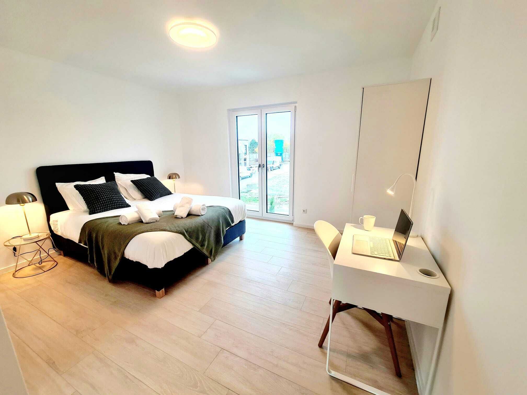 A modern bedroom with a bed, bedside table, lamp, and a desk with a laptop.
