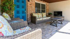 Ein überdachter Patio-Bereich mit Korbmöbeln, einer blauen Tür und einem Fernseher auf einem Standfuß.