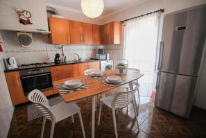 A compact kitchen with wooden cabinets, a stainless steel refrigerator, a dining table with white chairs, and a microwave. - Besugo's House Rooms & Apartment (Nazaré)