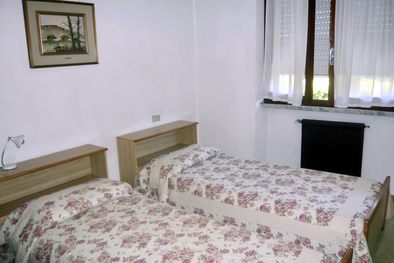 Ein Schlafzimmer mit zwei Einzelbetten, einem Holz-Kopfteil, einem Nachttisch mit Lampe, einem gerahmten Bild an der Wand und einem Fenster mit Vorhängen.