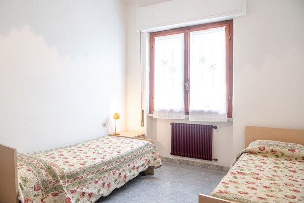 A small, well-lit room with two single beds, a radiator, a window with curtains, and a bedside lamp.