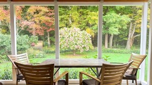 Un porxo cobert amb una taula i cadires de fusta, grans finestres que ofereixen vista a un jardí amb arbres i arbustos.
