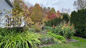 Un jardí amb diverses plantes i flors, una tanca blanca i una casa amb un porxo.