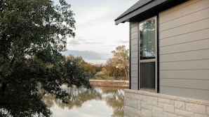 A house with a stone foundation and a window overlooking a calm body of water, surrounded by trees. - Ranch Sunsets - Luxury Lakeview Ranch Villa at Twin Canyons Longhorn Ranch (Granbury)