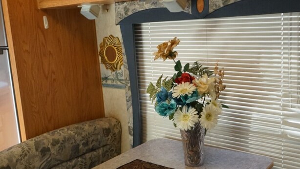A compact RV kitchen with wooden cabinets, a dining area with a table and bench, and a decorative flower arrangement on the windowsill. - Camper in Dandridge. Close to the Smoky Mountains and Dollywood  (Dandridge)