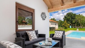 A sunlit outdoor seating area with wicker furniture and a glass coffee table, overlooking a swimming pool and a garden.