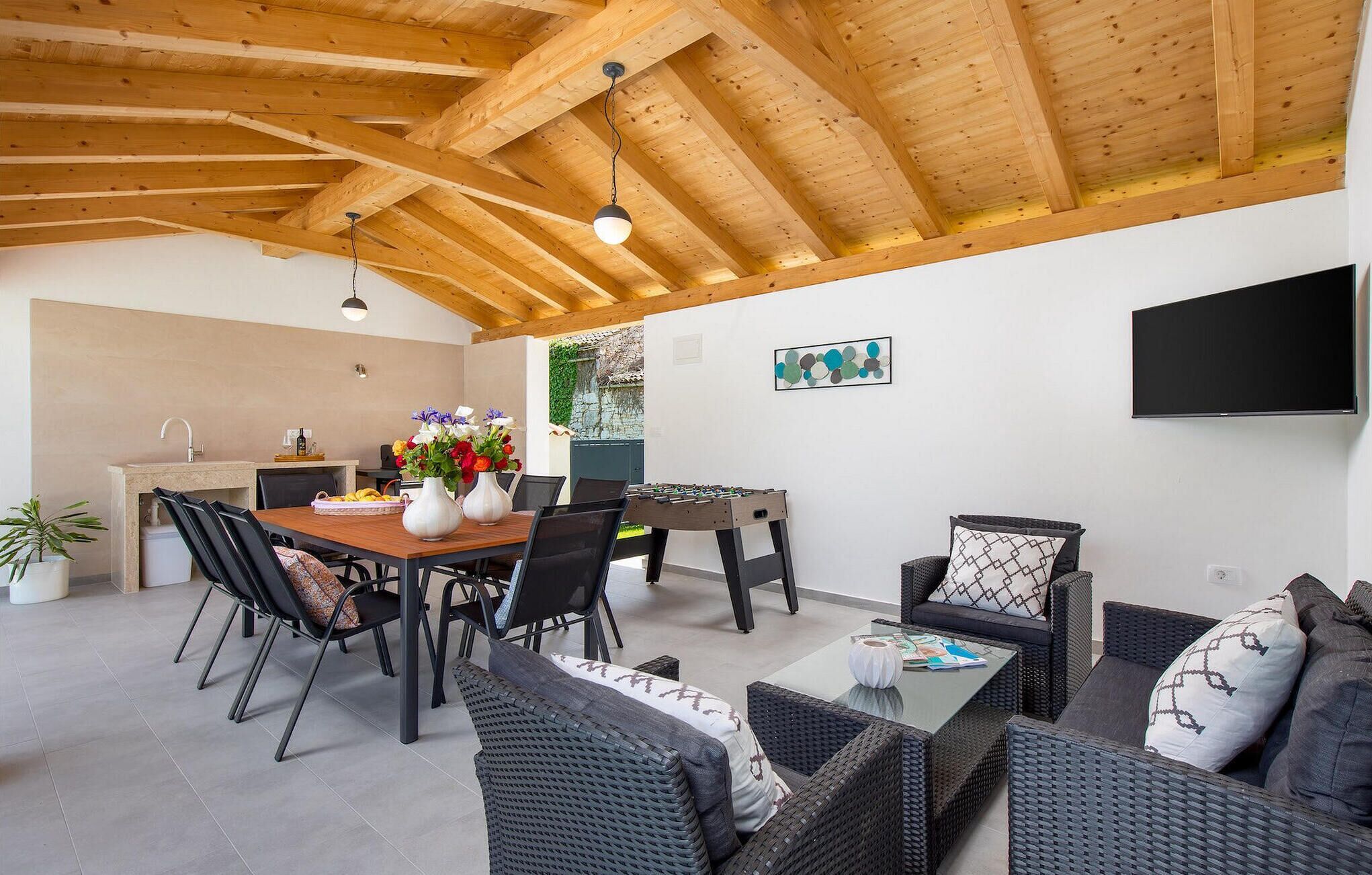 Un espace repas extérieur doté d’un plafond en bois, d’une table avec des chaises, d’un canapé et d’un téléviseur fixé au mur.
